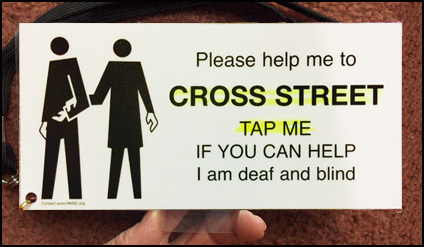 The card has a drawing of one person guiding another, and says: 'Please help me to -- CROSS STREET -- TAP ME -- IF YOU CAN HELP -- I am deaf and blind.' The words 'Cross street, tap me' are highlighted in yellow.  The card has small holes at the top right and bottom left; a neck cord attached to the bottom left corner.  The card is held by a see-through tab at the bottom.